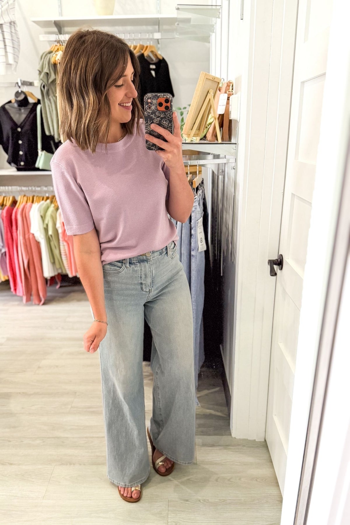 Woman taking a mirror selfie in a clothing store wearing a lavender short sleeve sweater and light blue jeans.
