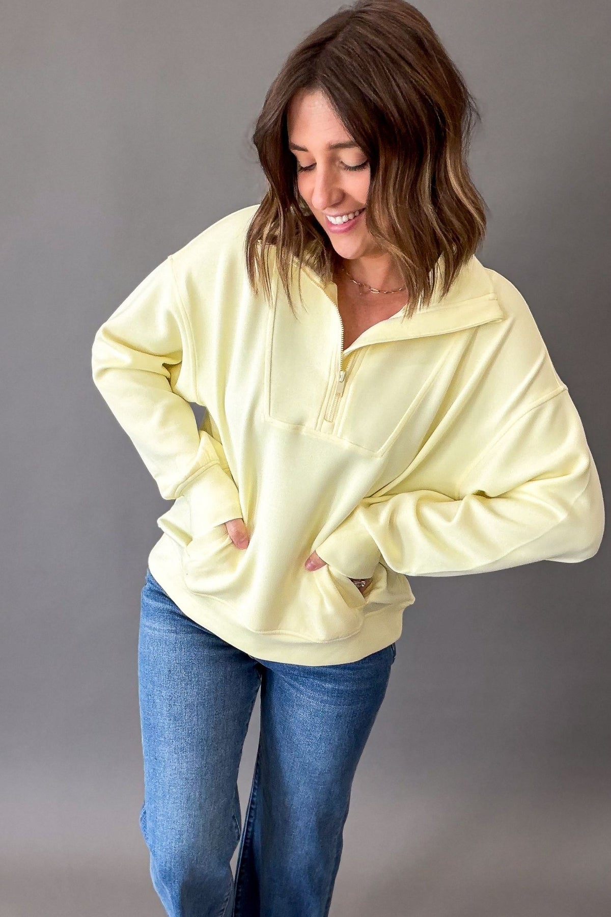 Woman wearing a light yellow oversized mock neck quarter zip and blue jeans against a gray background