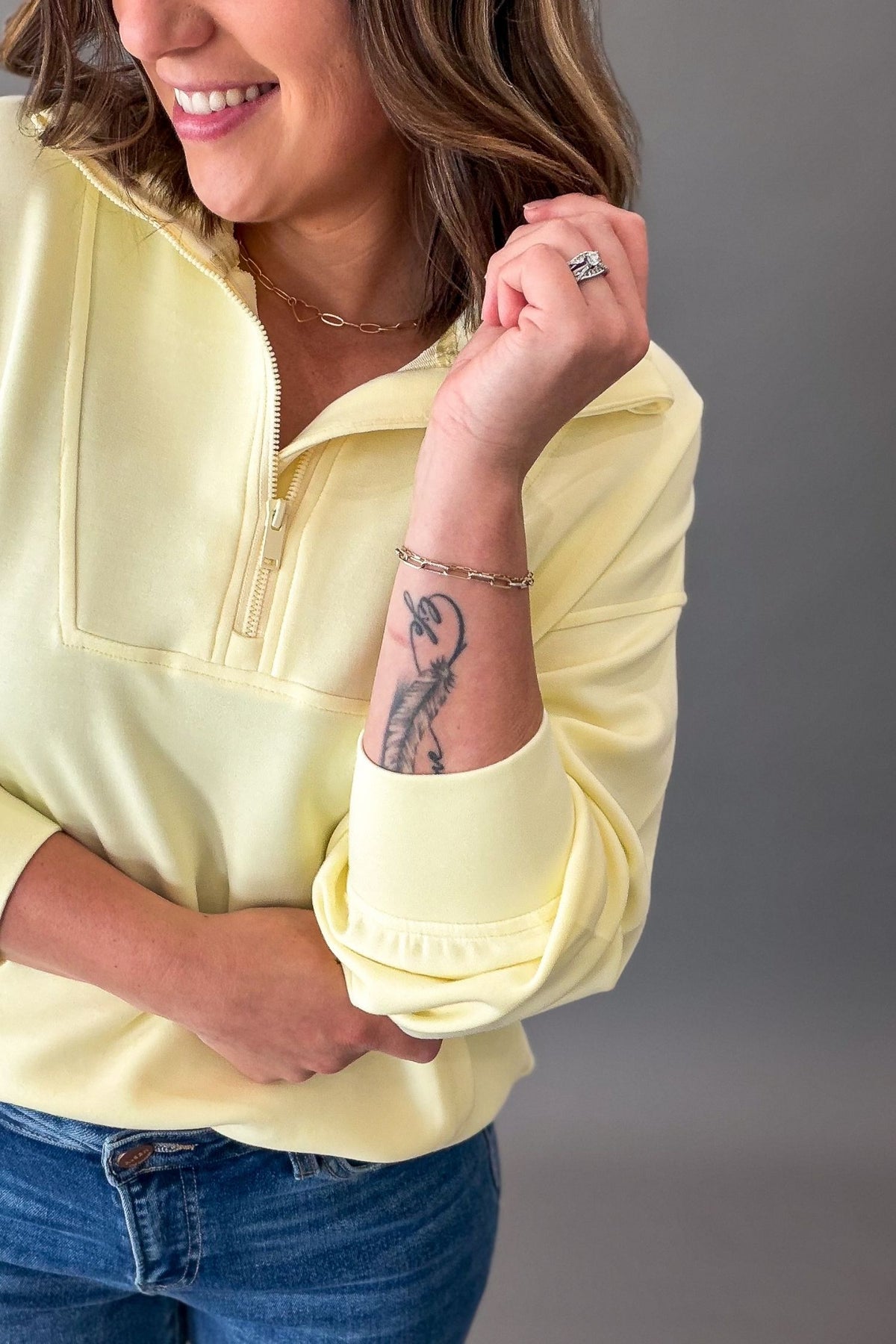 Woman wearing a light yellow mock neck quarter zip long-sleeve pullover with rolled-up sleeves, blue jeans, and a gray background.