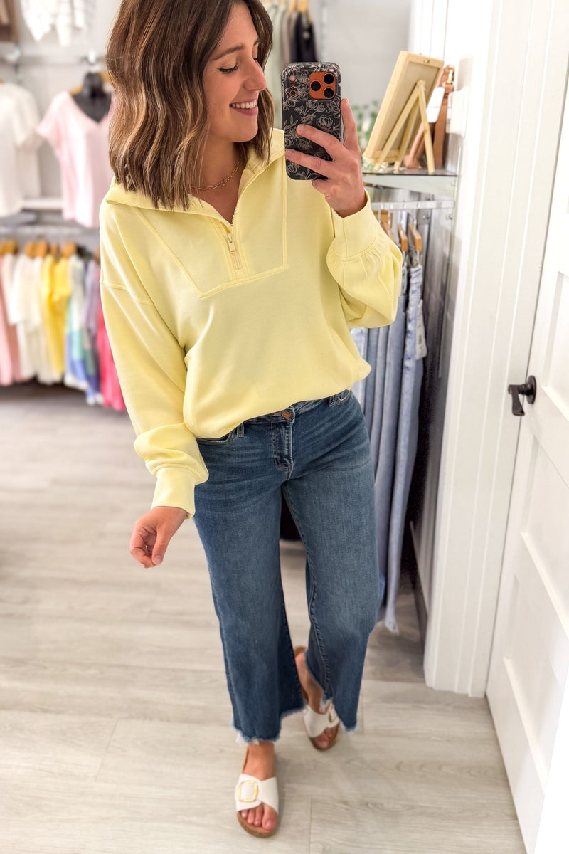 Woman taking a selfie wearing a light yellow oversized mock neck quarter zip and blue jeans against a gray background