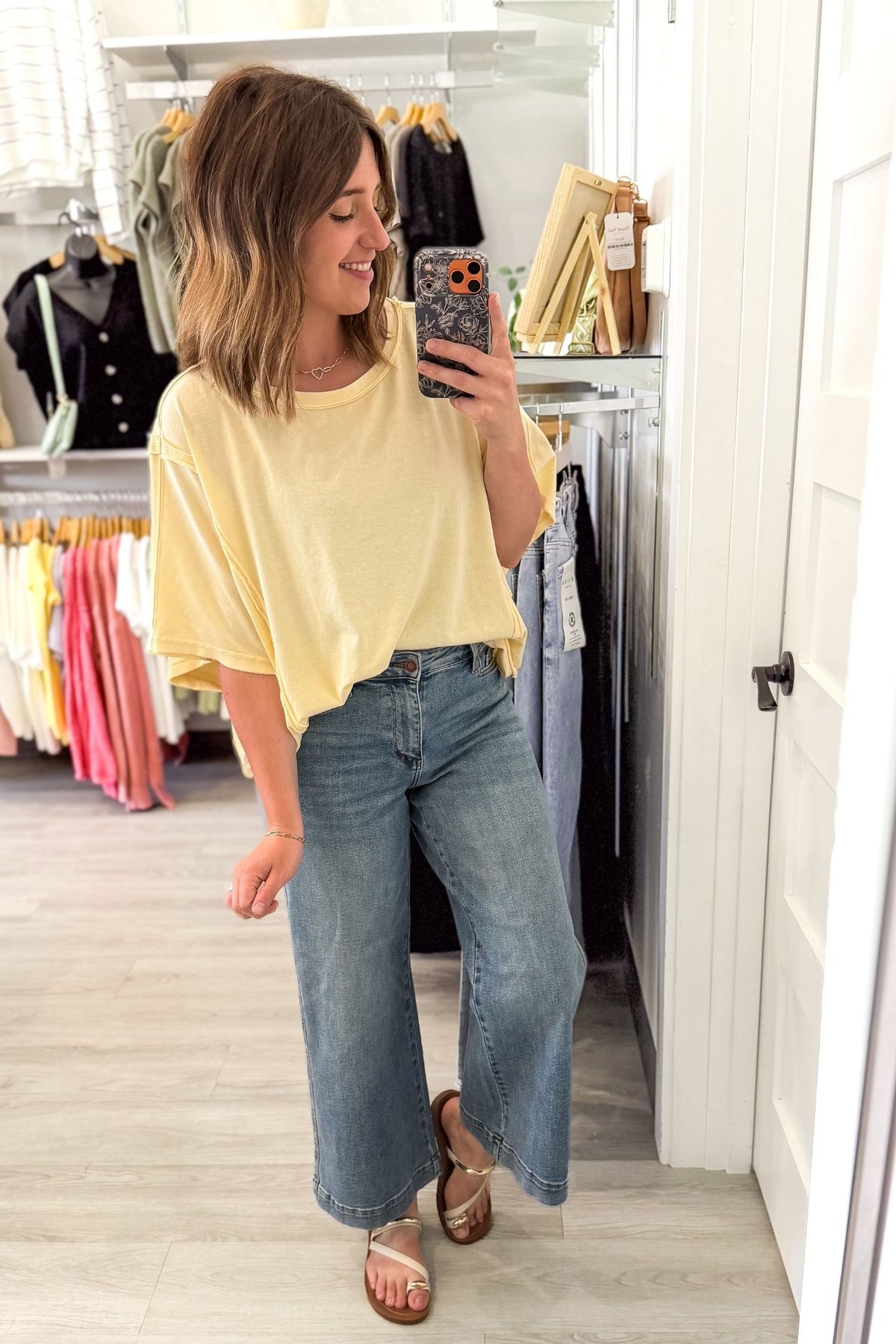 Woman taking a mirror selfie wearing a yellow top and blue jeans in a clothing store.