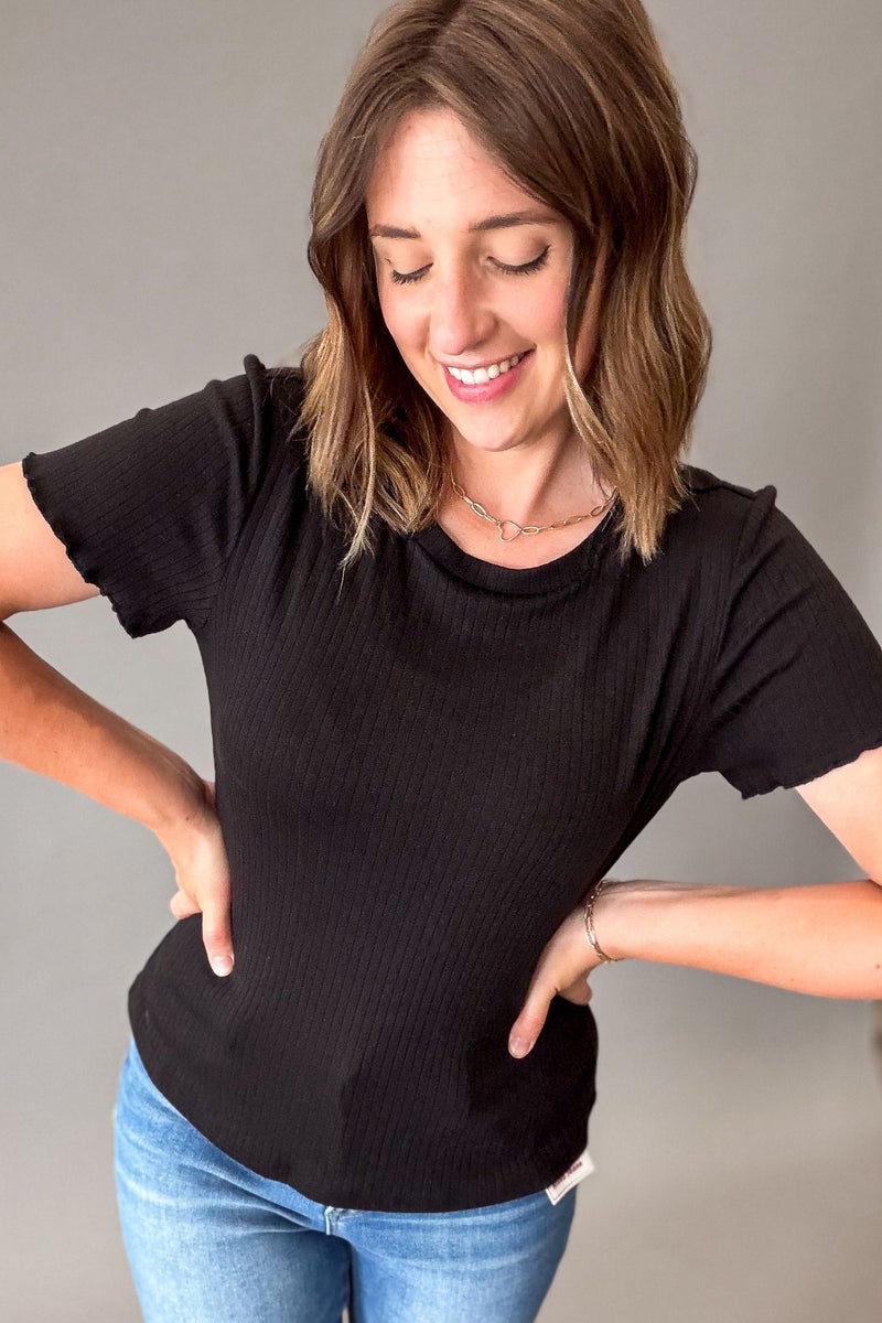 A woman wearing a black short-sleeve ribbed t-shirt and blue jeans, posing with her hands on her hips against a plain gray background.