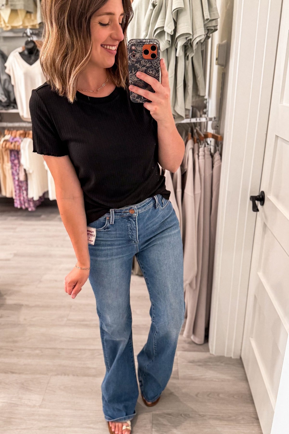 A woman takes a mirror selfie in a clothing store, wearing a black short-sleeve crewneck t-shirt and medium-wash blue flare jeans.