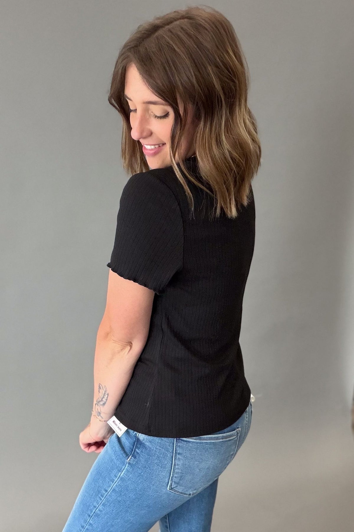 A woman with shoulder-length brown hair is shown from the back wearing a black short-sleeve ribbed top and light blue jeans against a plain gray studio background.