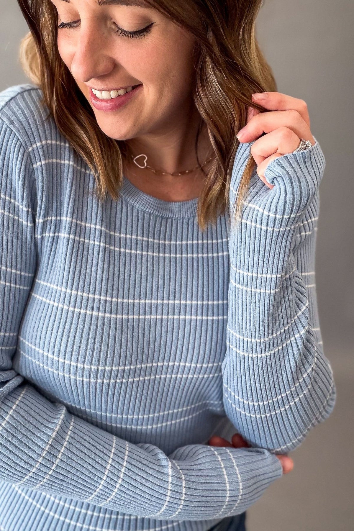 A woman wearing a light blue ribbed knit sweater with thin white horizontal stripes, shown in a close-up shot against a neutral gray background.