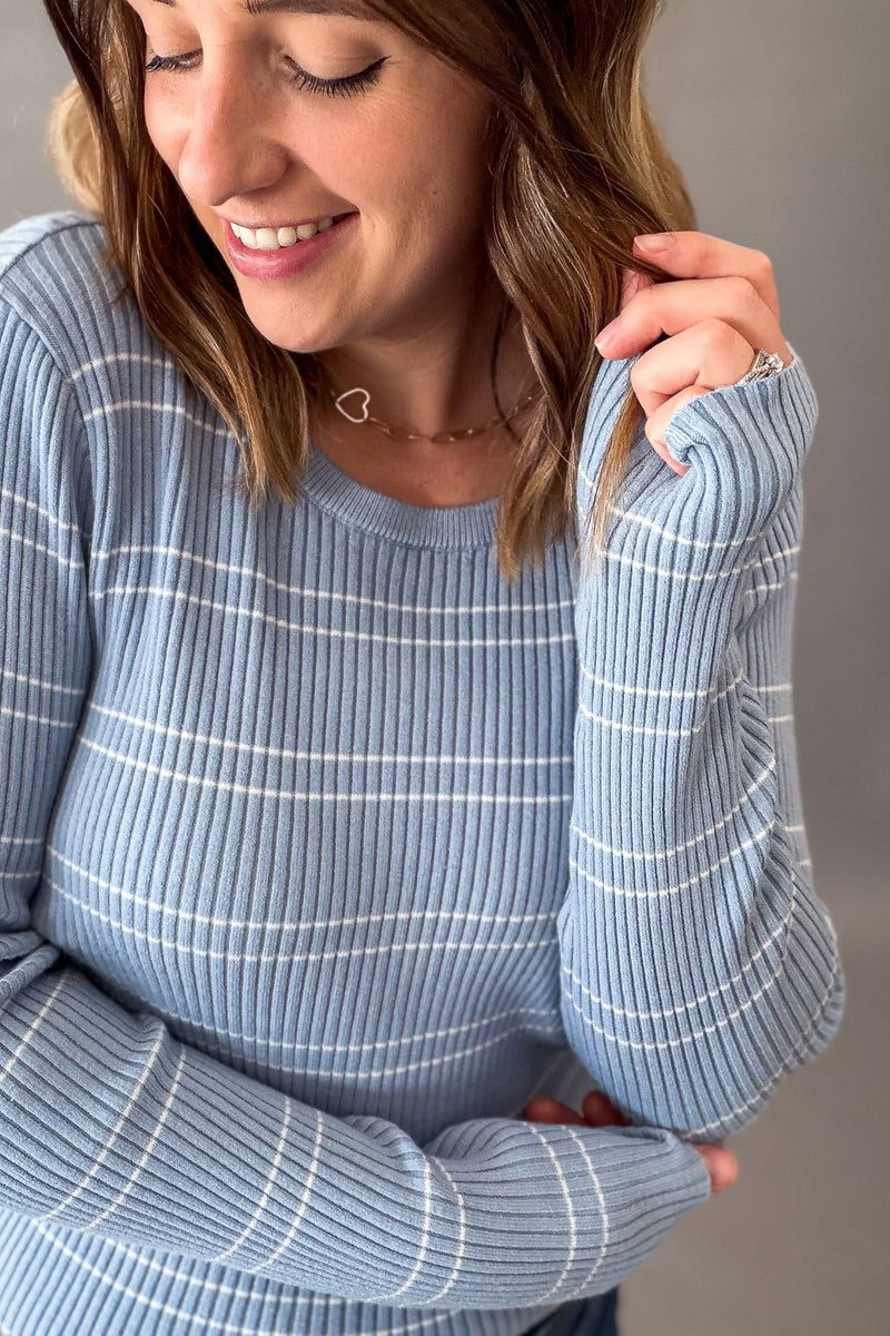 A woman wearing a light blue ribbed knit sweater with thin white horizontal stripes, shown in a close-up shot against a neutral gray background.