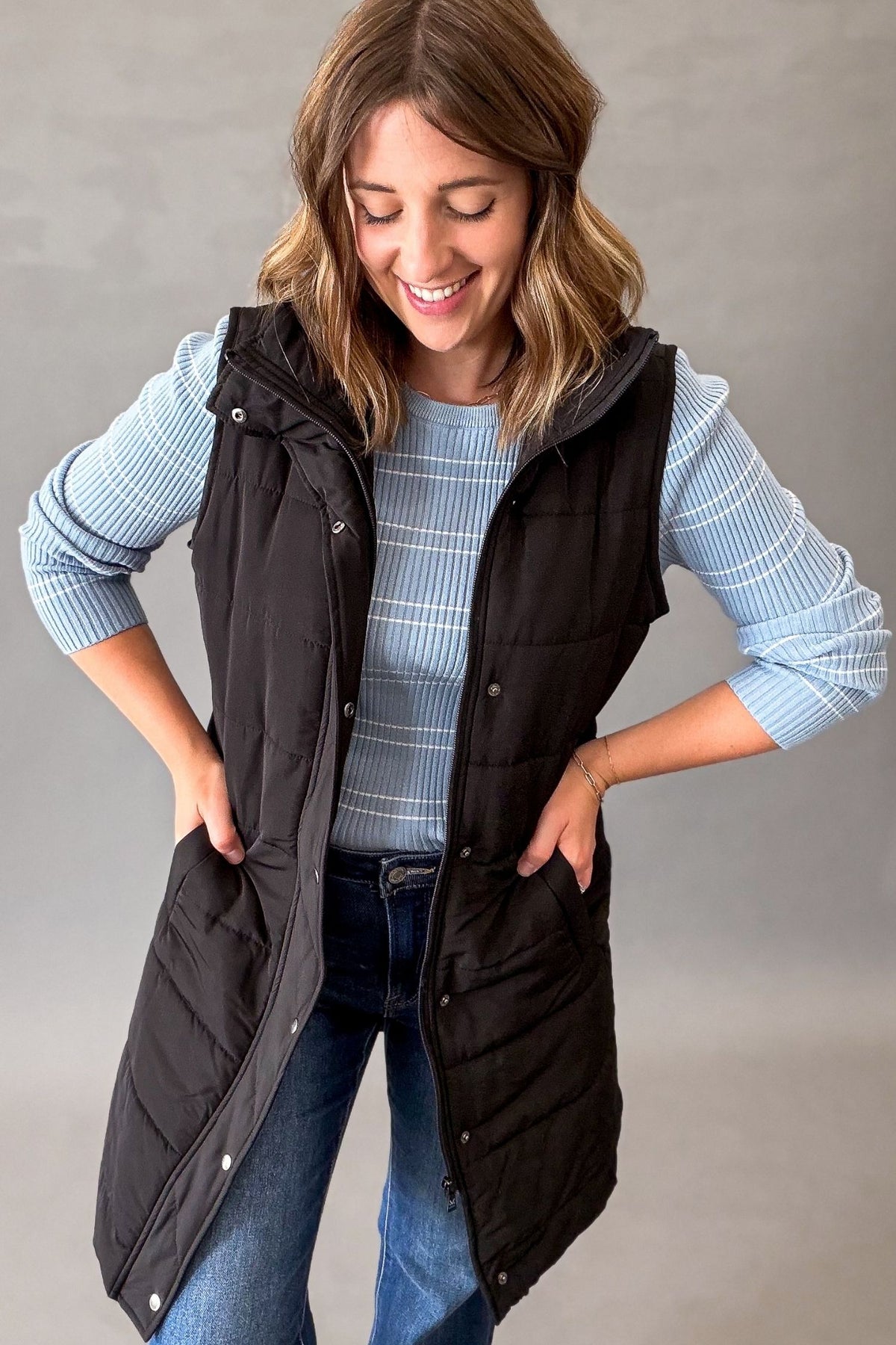 A woman wearing a black sleeveless puffer vest over a light blue and white striped long-sleeve shirt, paired with blue jeans, posing against a neutral gray studio background.