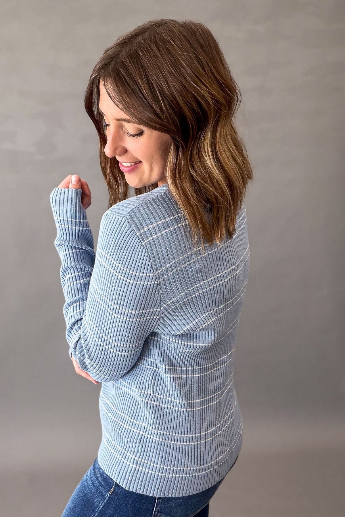 A woman with shoulder-length brown hair is shown from the back wearing a light blue and white striped ribbed knit sweater and blue jeans, standing against a neutral gray studio background.