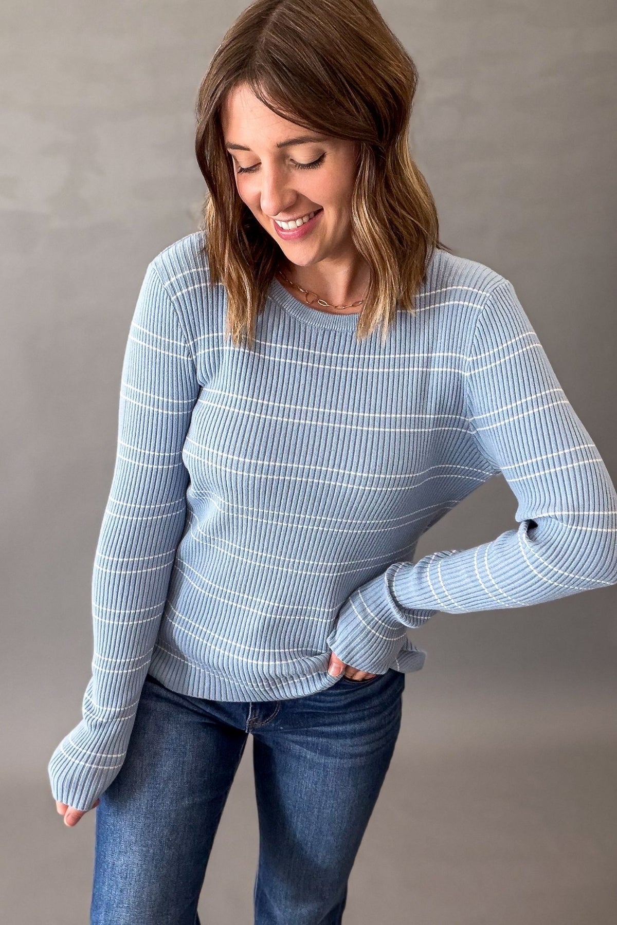 A woman wearing a light blue and white striped ribbed knit sweater with fingerless sleeves, paired with blue jeans, standing against a neutral gray wall.