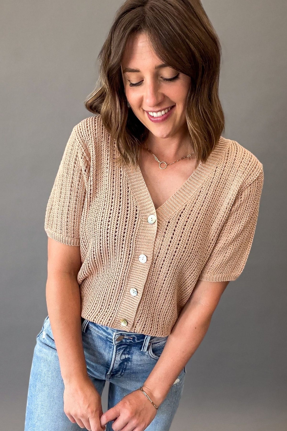 woman wearing a Light mocha short sleeve knit sweater with a V-neckline, functional button front, open-knit texture, and a soft, stretchy fit styled with light wash denim.