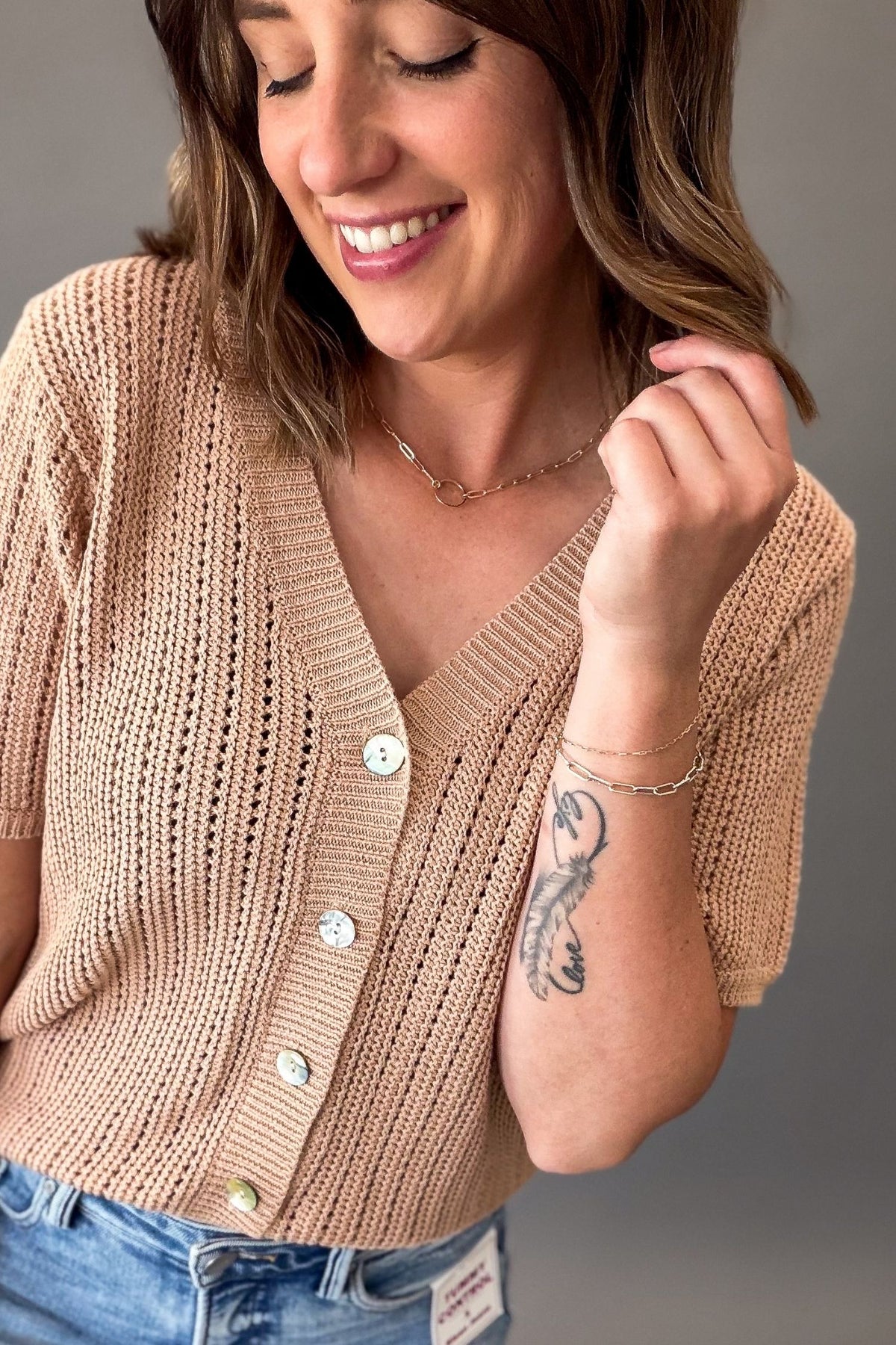 woman wearing a Light mocha short sleeve knit sweater with a V-neckline, functional button front, open-knit texture, and a soft, stretchy fit styled with light wash denim.