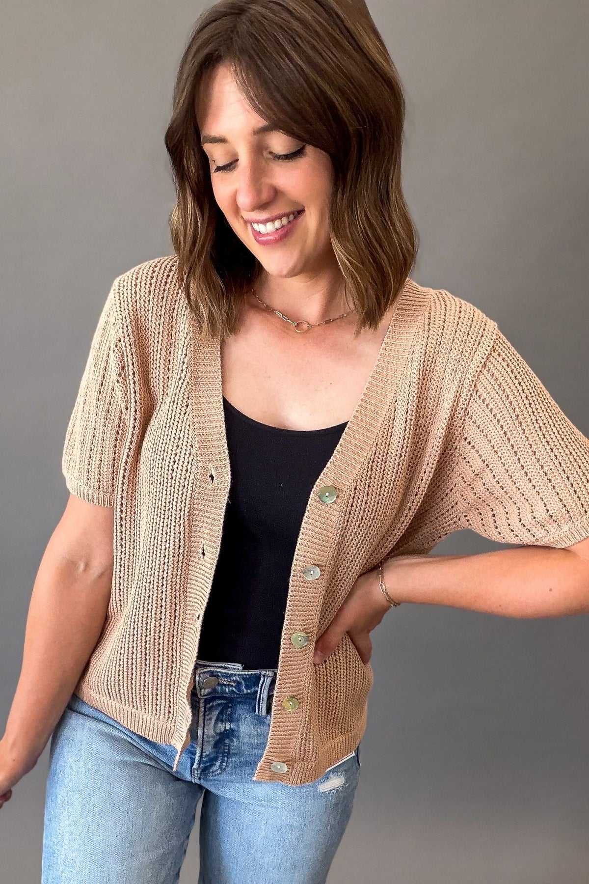 woman wearing a Light mocha short sleeve knit sweater open with a V-neckline, functional button front, open-knit texture, and a soft, stretchy fit styled with light wash denim.