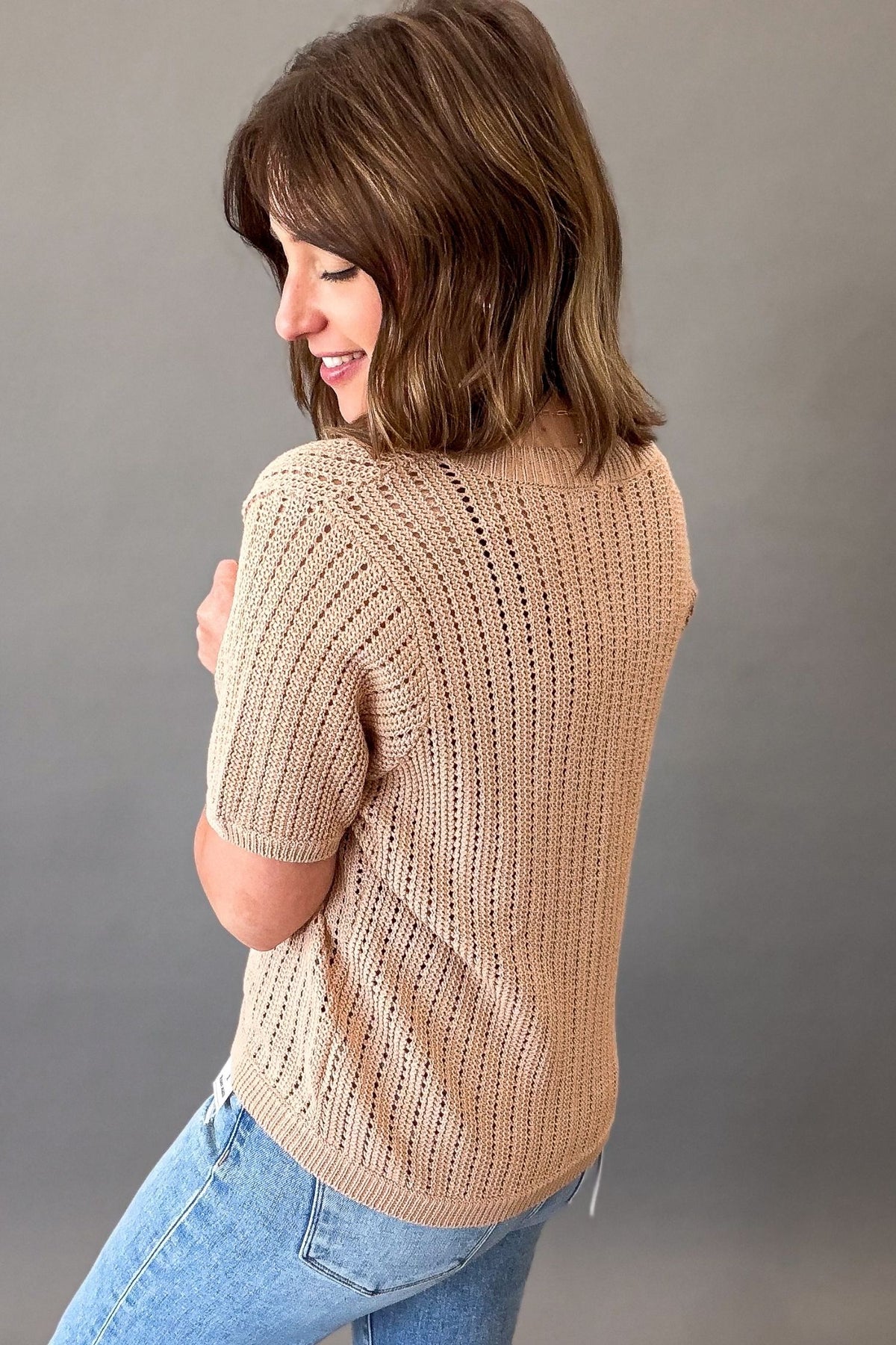 woman wearing a Light mocha short sleeve knit sweater with a V-neckline, functional button front, open-knit texture, and a soft, stretchy fit styled with light wash denim showing the back and side details