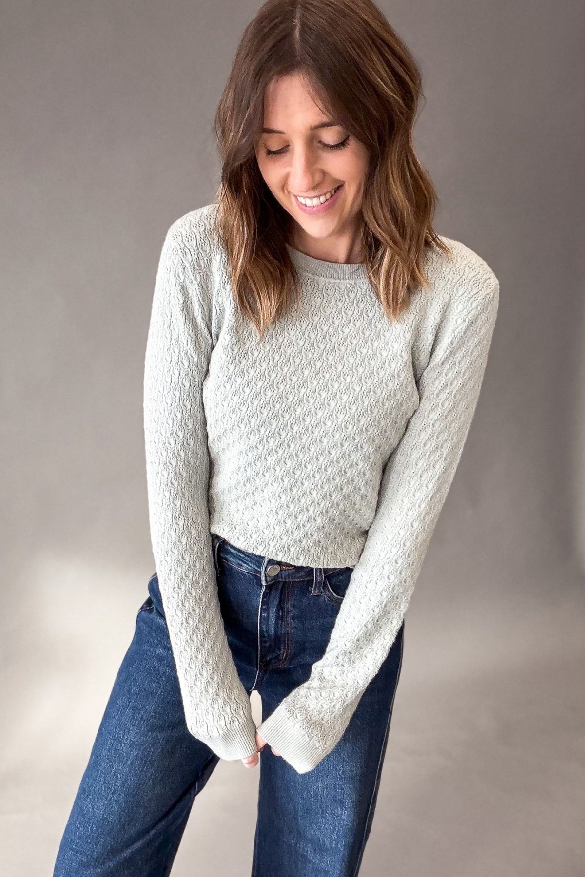 A woman wearing a light sage textured knit sweater and blue jeans, standing against a neutral gray studio background.