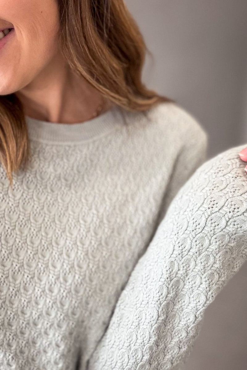 Close-up of a woman wearing a light sage textured knit sweater, showing the detailed lace-like pattern on the sleeve and chest area.