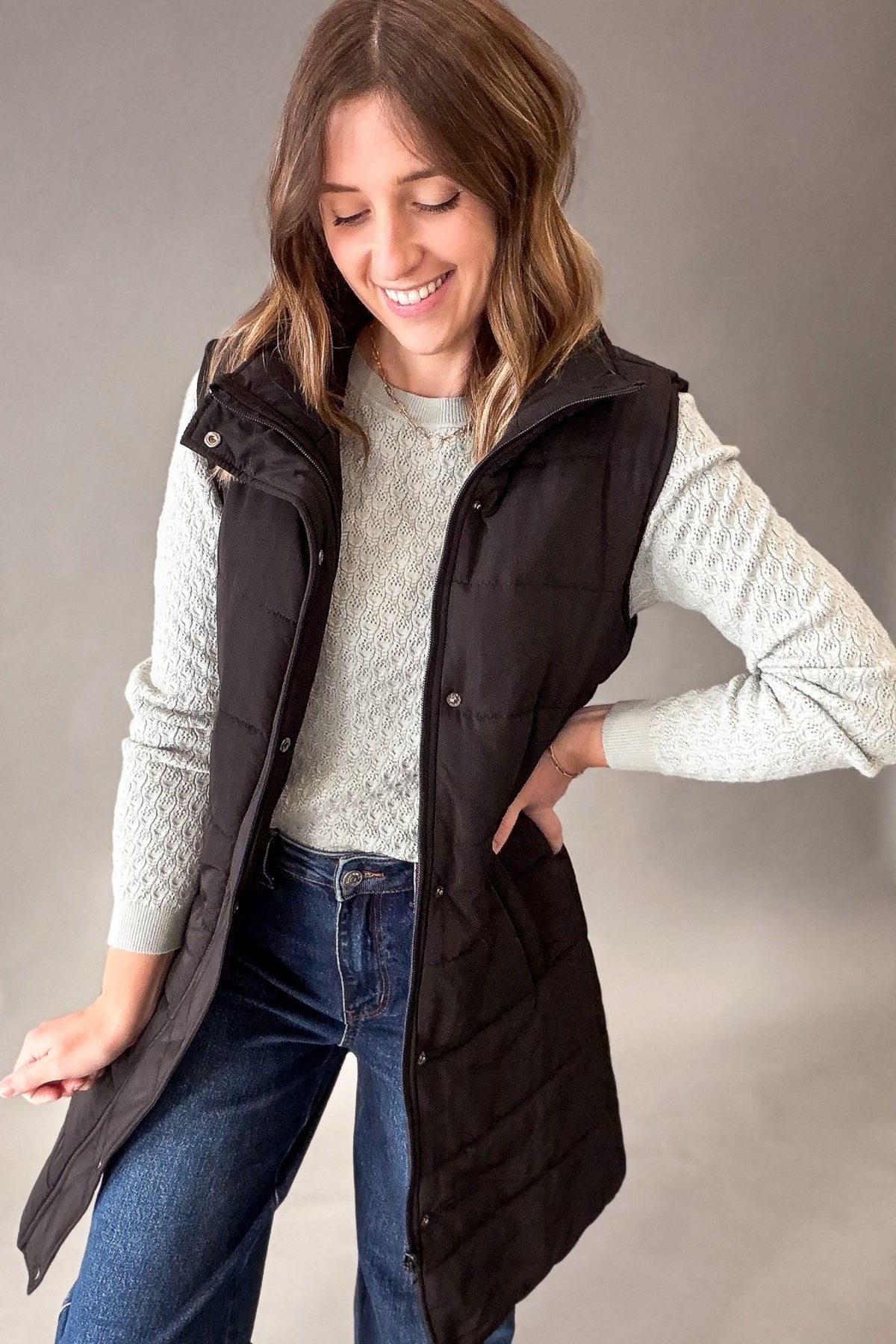 A woman wearing a black sleeveless puffer vest over a light sage knit sweater and blue jeans, standing against a plain gray studio background.