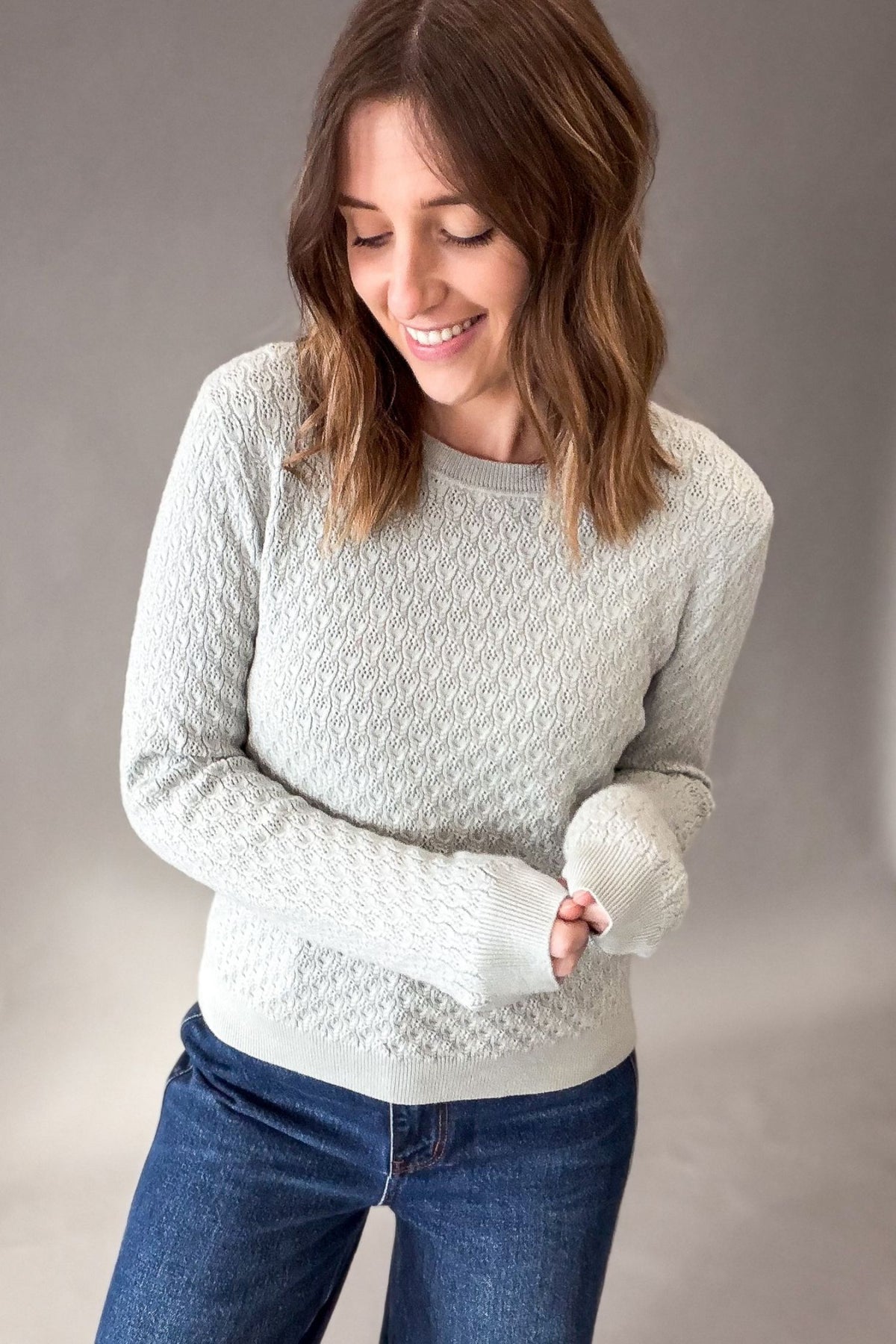 A woman wearing a light sage textured knit sweater and blue jeans, standing against a neutral gray studio background.