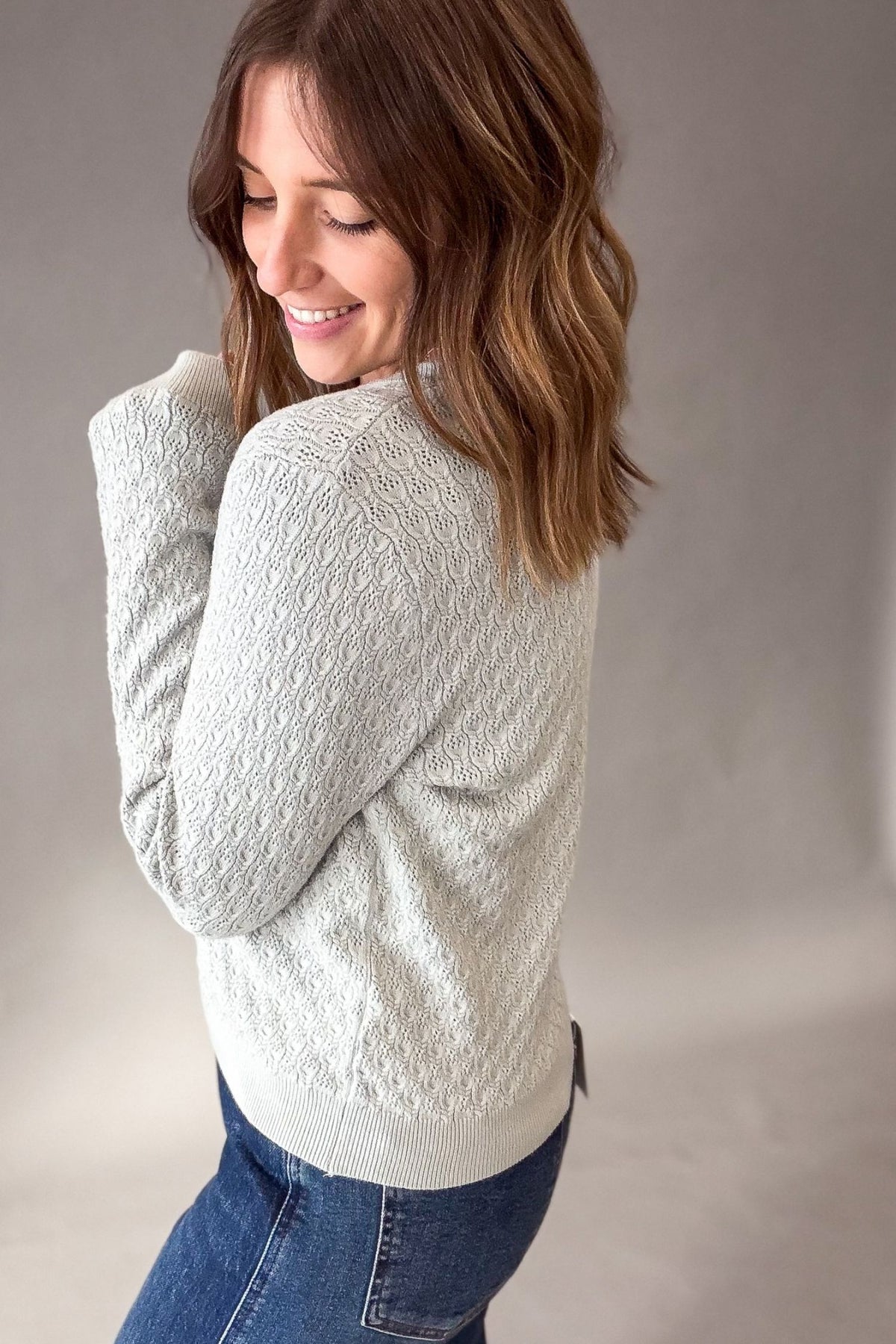 A woman wearing a light sage cable-knit sweater and blue jeans, shown from a side-back angle against a neutral gray studio background.