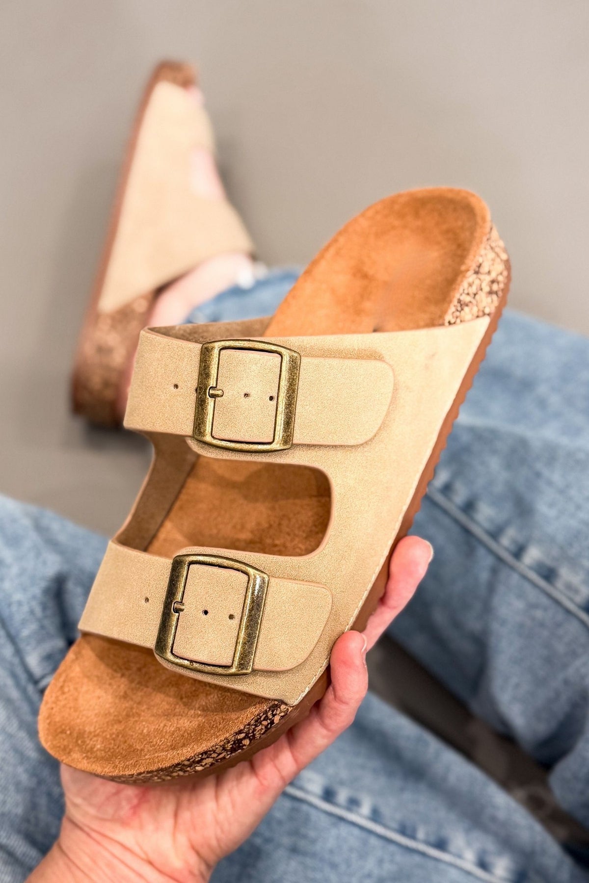 A person holds a taupe double-strap slide sandal with gold-tone buckles and a cork-look sole. Another matching sandal is visible in the background on blue jeans against a neutral gray wall.