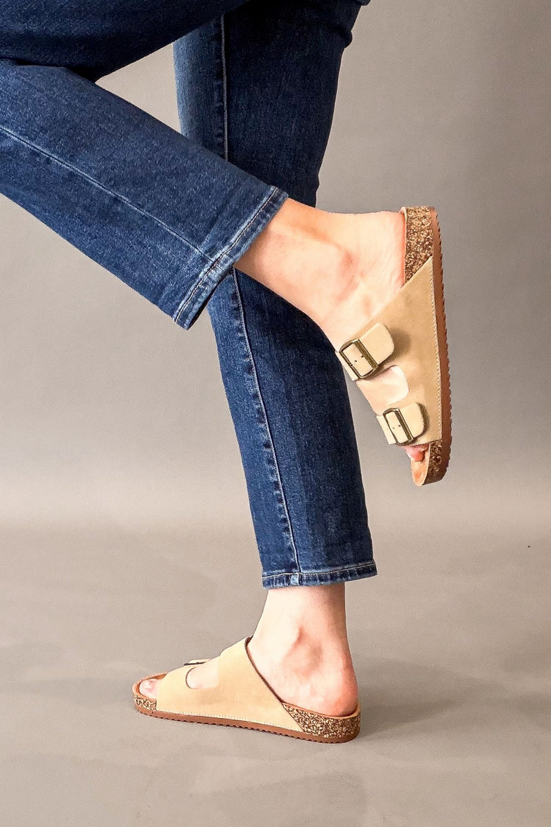 A person wearing taupe double-strap slide sandals with cork-look soles and gold-tone buckles, paired with blue jeans, shown against a neutral gray studio background.