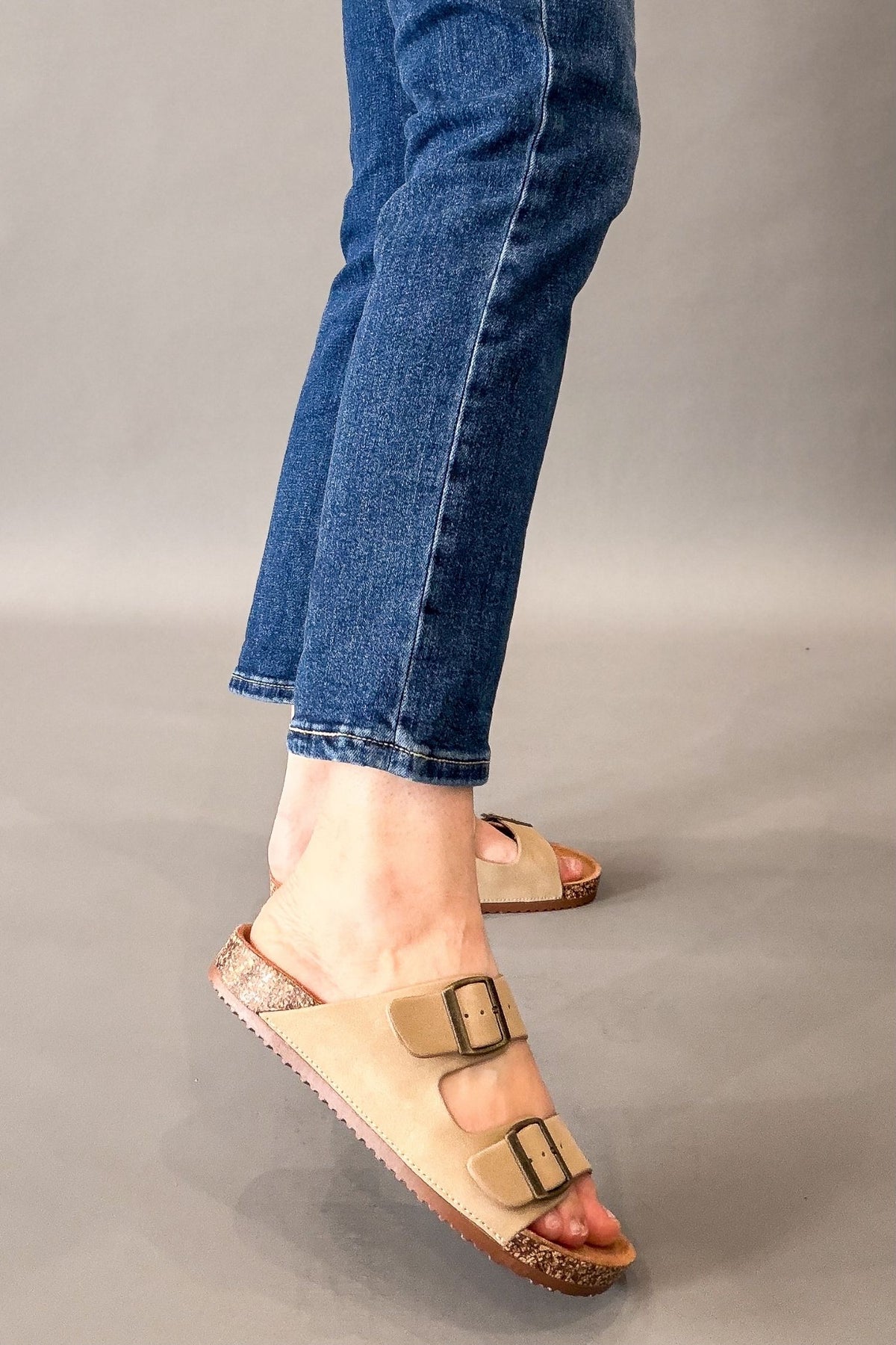 A person wearing taupe double-strap slide sandals with brown cork-look soles and blue jeans, shown from the knees down against a plain gray studio background.