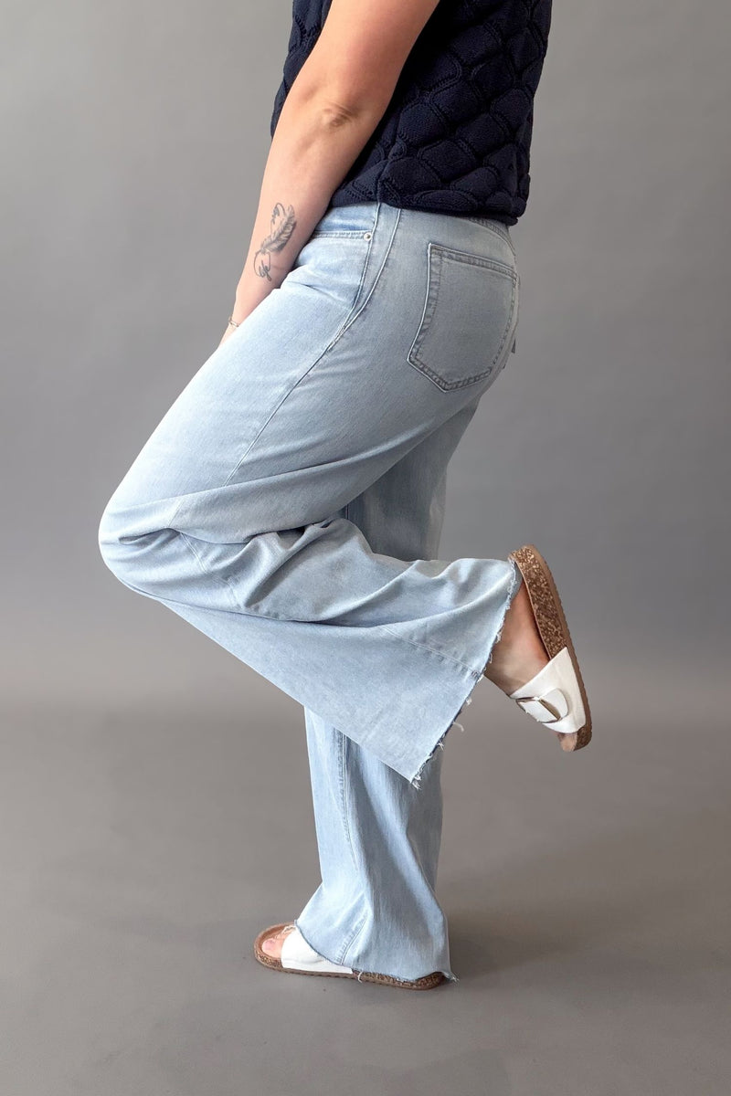 A woman wearing light-wash wide-leg Mica jeans with a raw hem, paired with a dark quilted top and white sandals, shown from the side against a plain gray studio background.