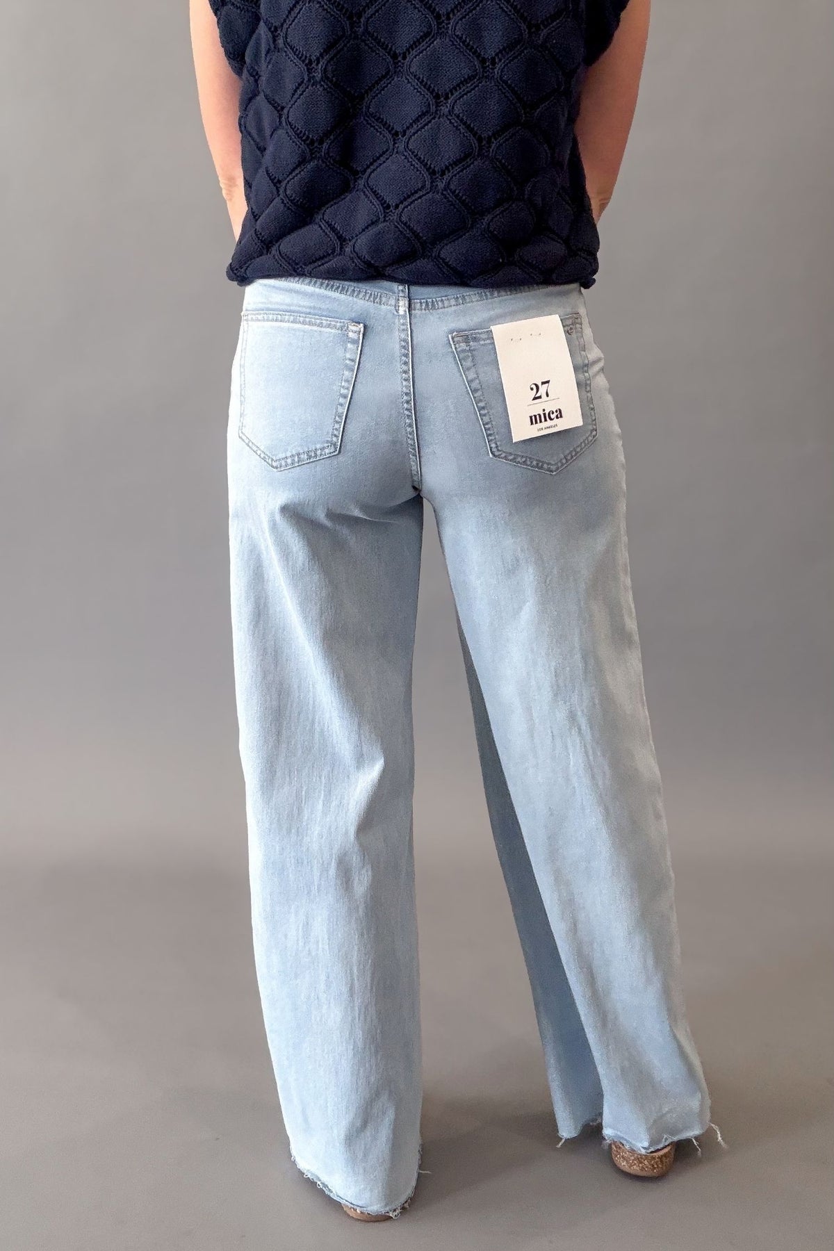 A person shown from the back wearing light-wash wide-leg jeans and a dark quilted top, standing against a plain gray studio background. A white paper tag is visible in the back pocket of the jeans.