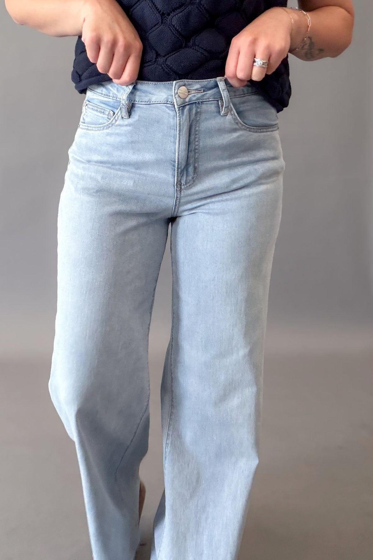 A person wearing light-wash blue straight-leg jeans and a dark navy quilted top, standing against a neutral gray studio background.