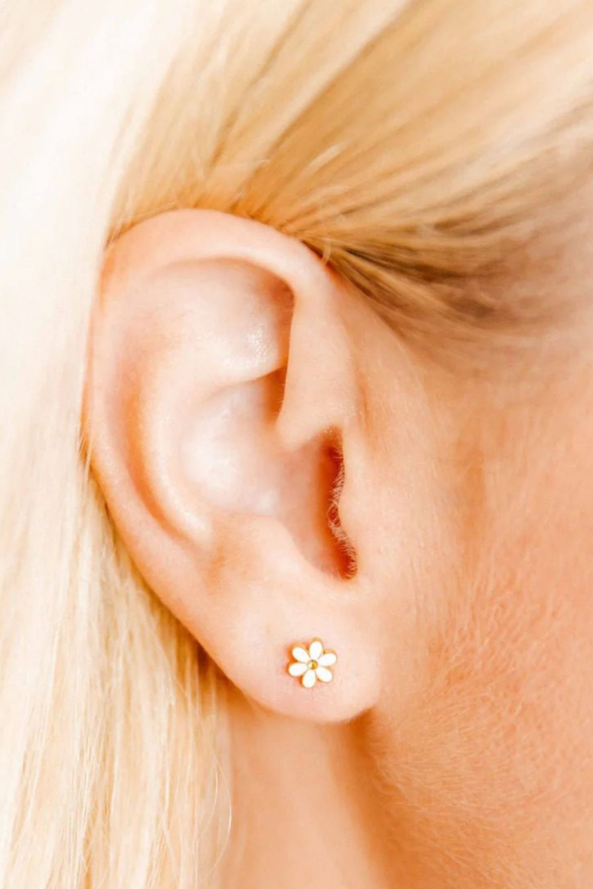 Close-up of an ear wearing a gold +white flower-shaped stud earring with blonde hair.