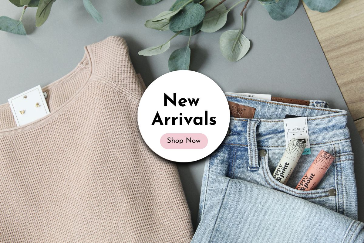 New arrivals clothing with a 'Shop Now' button on a gray surface with green leaves.
