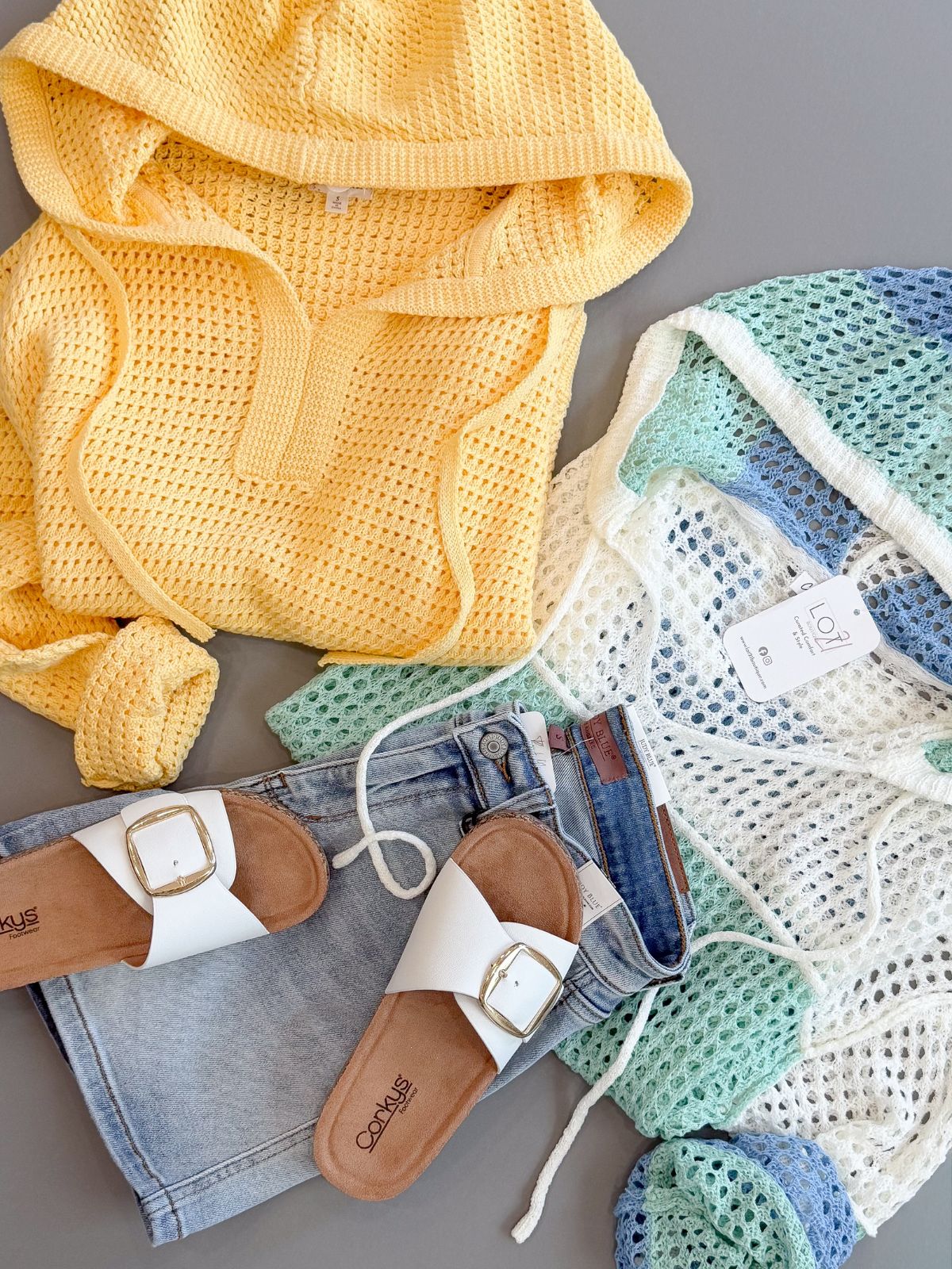 Yellow textured fabric, denim shorts, white sandals with brown soles, and a green textured fabric on a gray surface.
