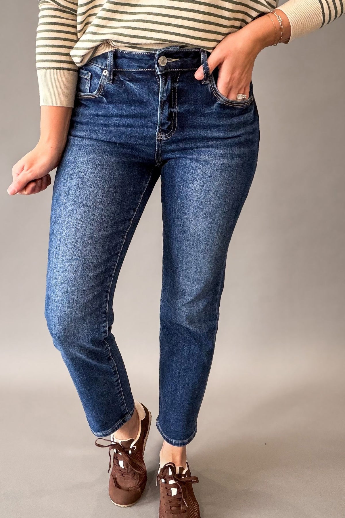 A person wearing MICA dark blue sMid rise straight crop jeans with a striped cream and brown top, brown boat shoes, and a bracelet, standing against a neutral gray studio background.