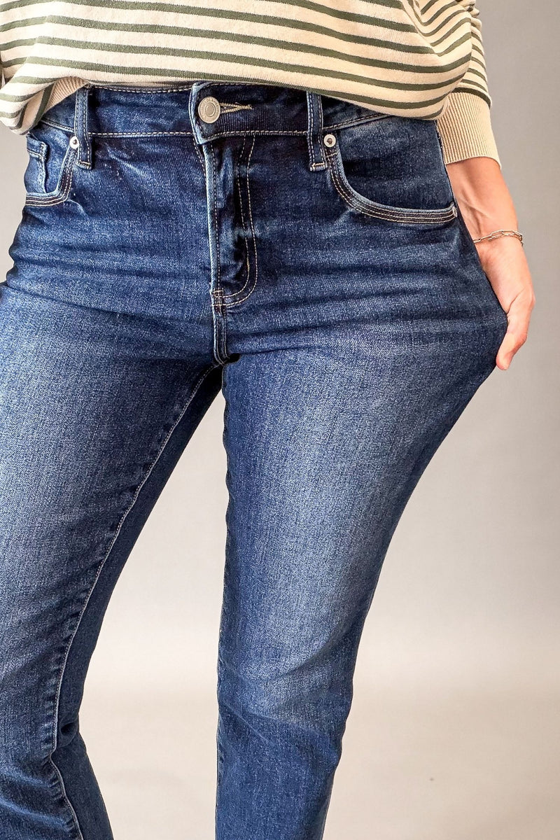 A close-up of a person wearing dark blue denim jeans with a striped beige and white top visible at the waist, photographed against a plain light gray studio background.