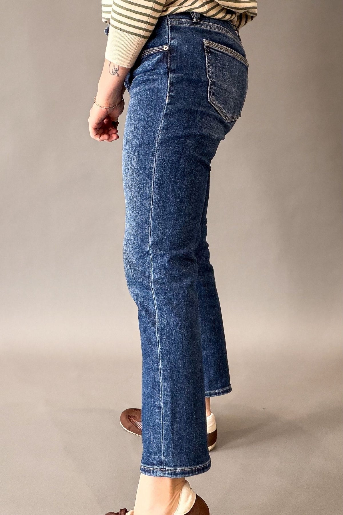 A person shown from the waist down wearing blue cropped straight jeans, a beige and white striped long-sleeve top, and brown shoes, standing against a neutral gray studio background.