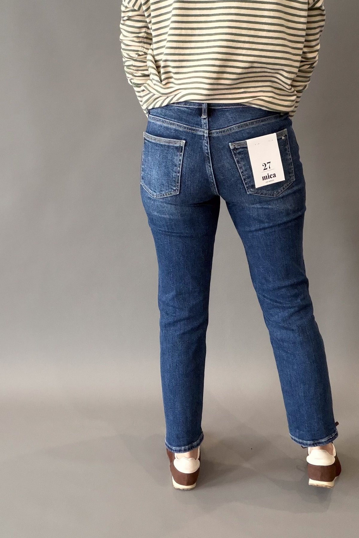 A person shown from the back wearing blue denim jeans with a striped cream and navy top, standing against a plain gray studio background. A white paper tag is visible in the back pocket.