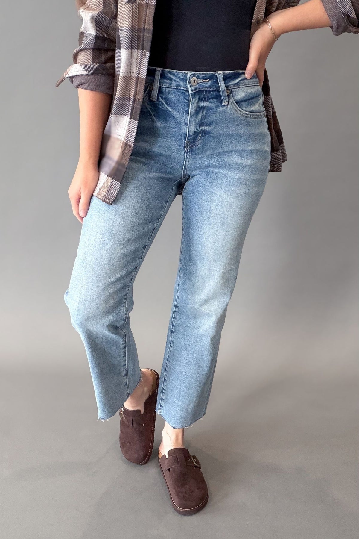 A person wearing light-wash blue cropped jeans with a black top, a brown plaid shirt draped off the shoulders, and brown slip-on shoes, standing against a plain gray studio background.
