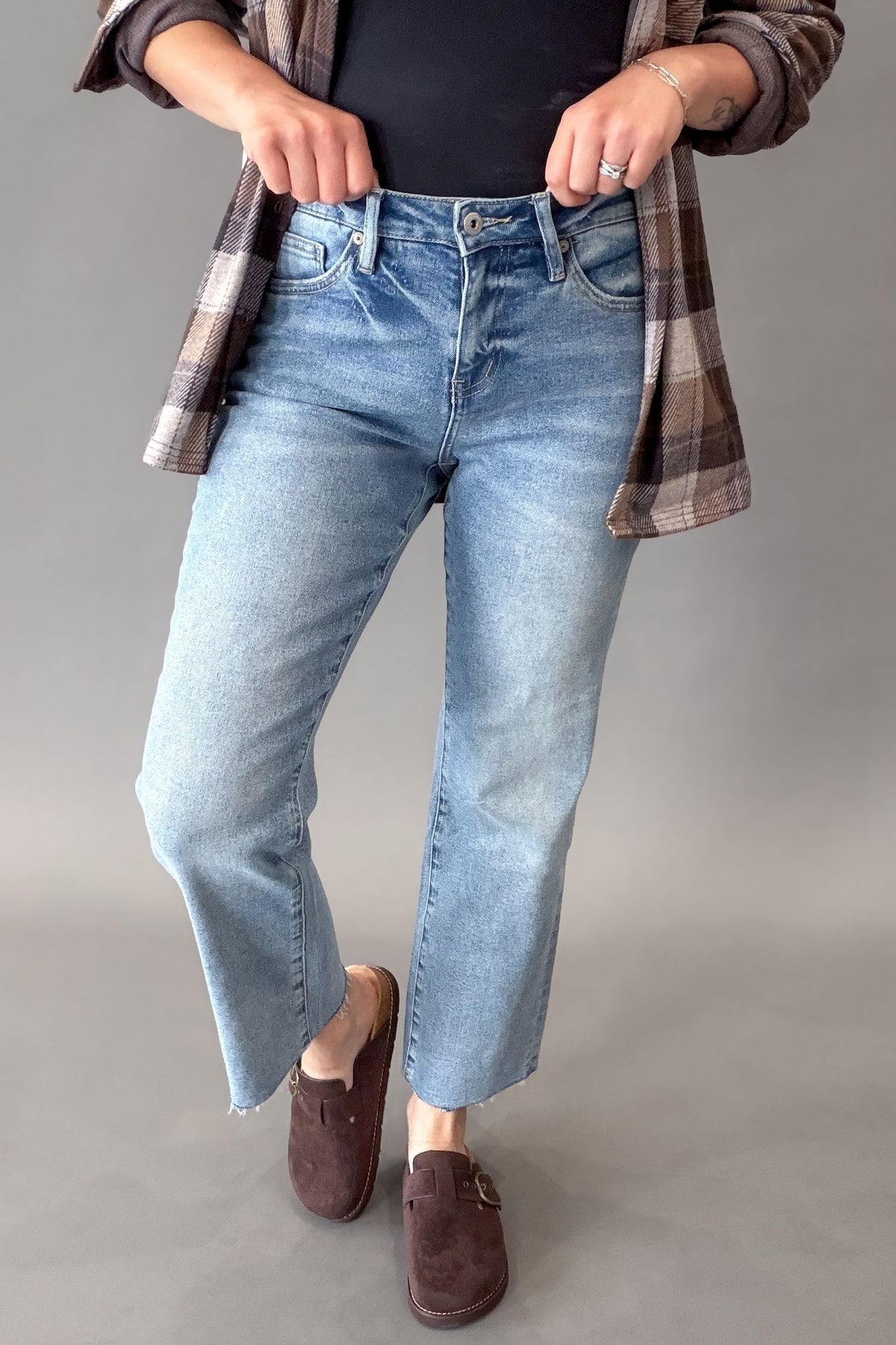 A person wearing light-wash blue cropped jeans with a brown plaid flannel shirt and dark brown suede loafers, standing against a plain gray studio background.