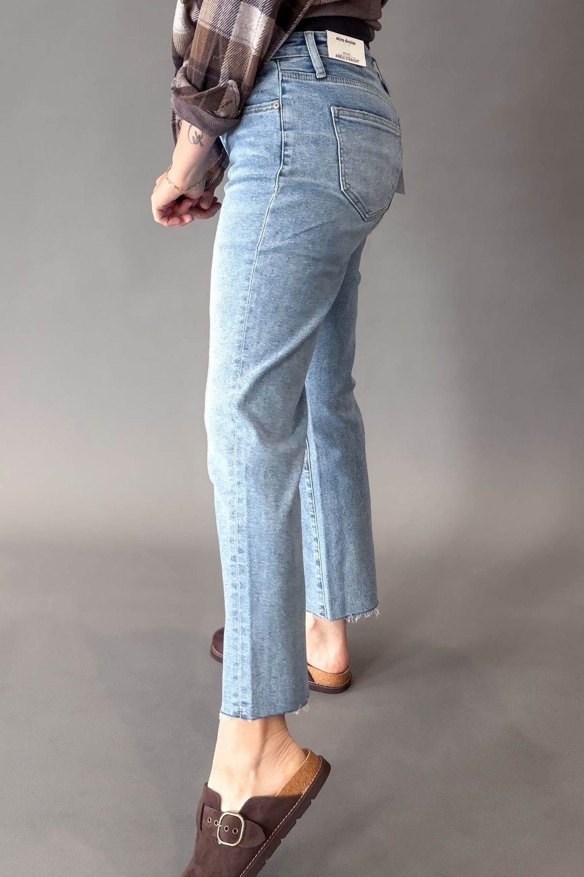 A person shown from the back wearing light-wash cropped denim jeans with a straight leg and raw hem, paired with brown mule loafers and a brown plaid shirt, against a neutral gray studio background.