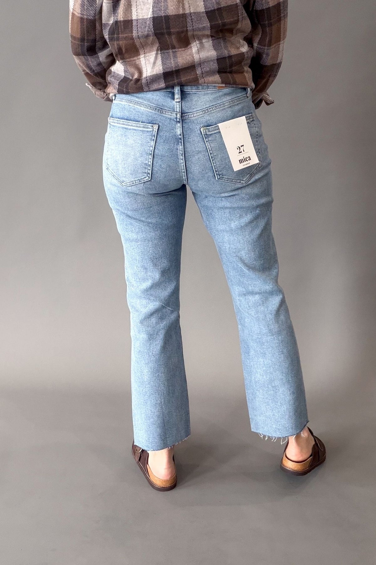 A person shown from the back wearing light-wash blue jeans with a brown and beige plaid shirt and brown sandals, standing against a plain gray studio background. A white paper tag is visible on the back pocket of the jeans.