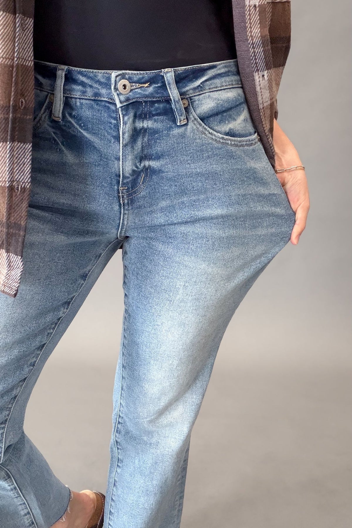 A close-up of a person wearing light-wash blue denim jeans, with a black top, a brown plaid overshirt, and a bracelet visible. The person is standing against a plain gray studio background.