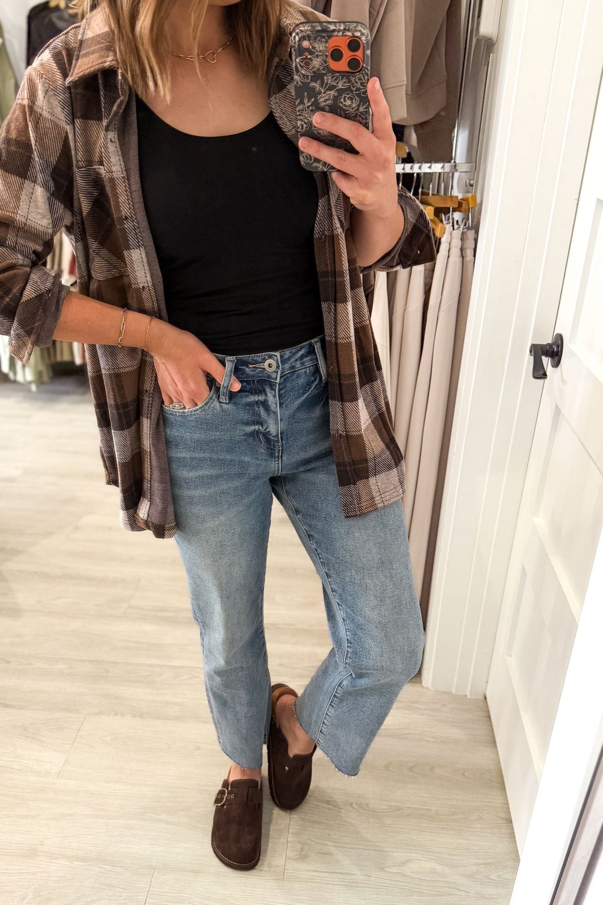 A person taking a mirror selfie in a clothing store, wearing a brown and beige plaid flannel shirt over a black tank top, light-wash blue jeans, and dark brown suede loafers.