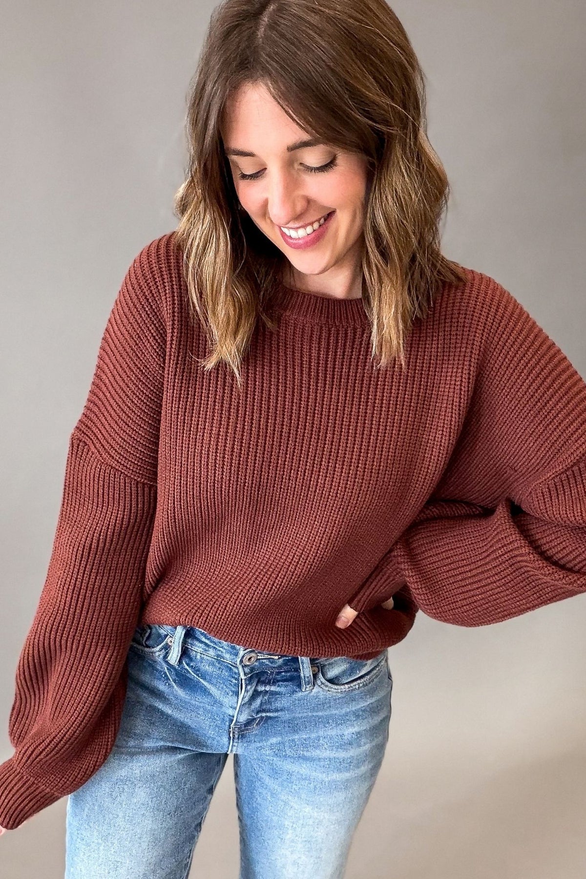 A woman wearing a maple brown-colored ribbed knit sweater and blue jeans, smiling and looking down against a plain gray studio background.