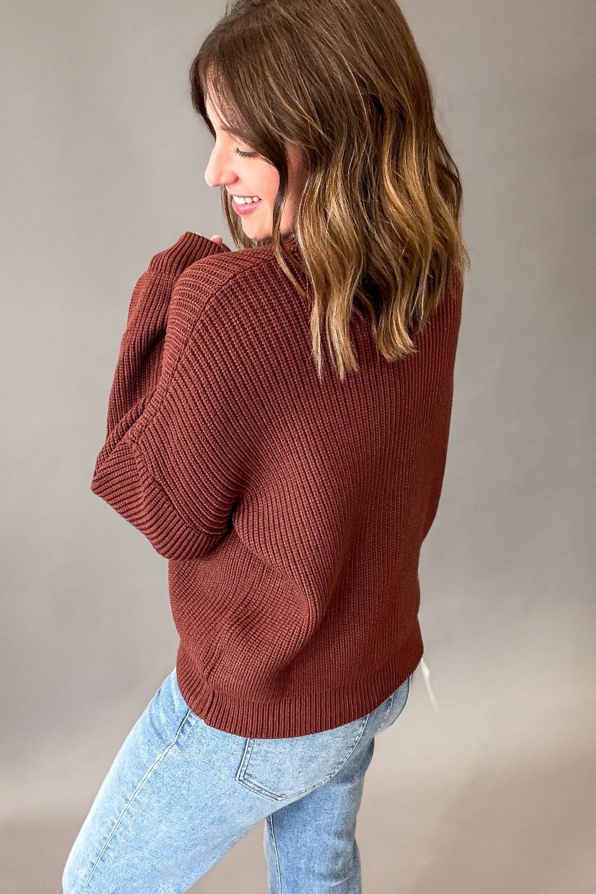 A woman wearing a maple-brown ribbed knit sweater and light blue jeans, photographed from the side against a plain gray studio background.