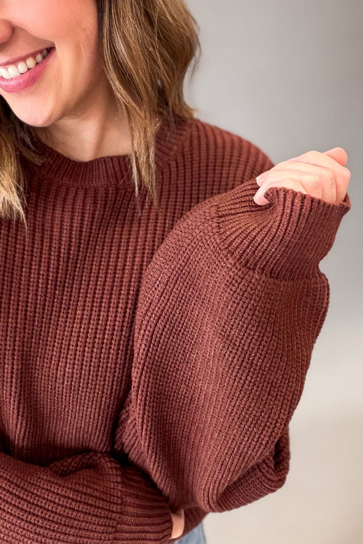A close-up of a woman wearing a ribbed knit sweater in a warm maple brown color, showing the texture of the fabric and the sleeve detail.