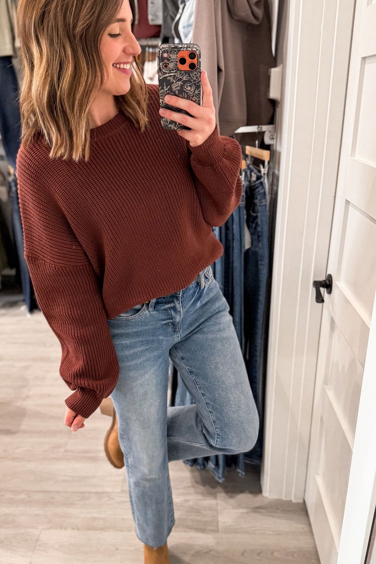 A woman takes a mirror selfie in a boutique, wearing a ribbed brown long-sleeve crop sweater and light-wash blue jeans.