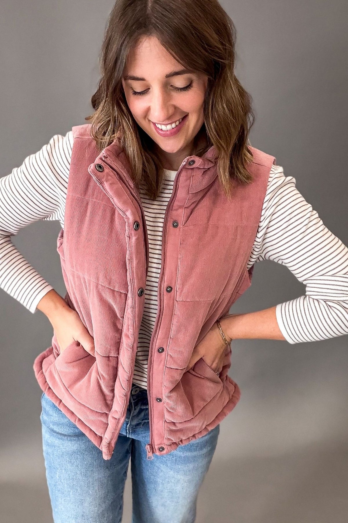 A woman wearing a mauve corduroy puffer vest over a white and gray striped long-sleeve shirt and blue jeans, standing against a neutral gray studio background.