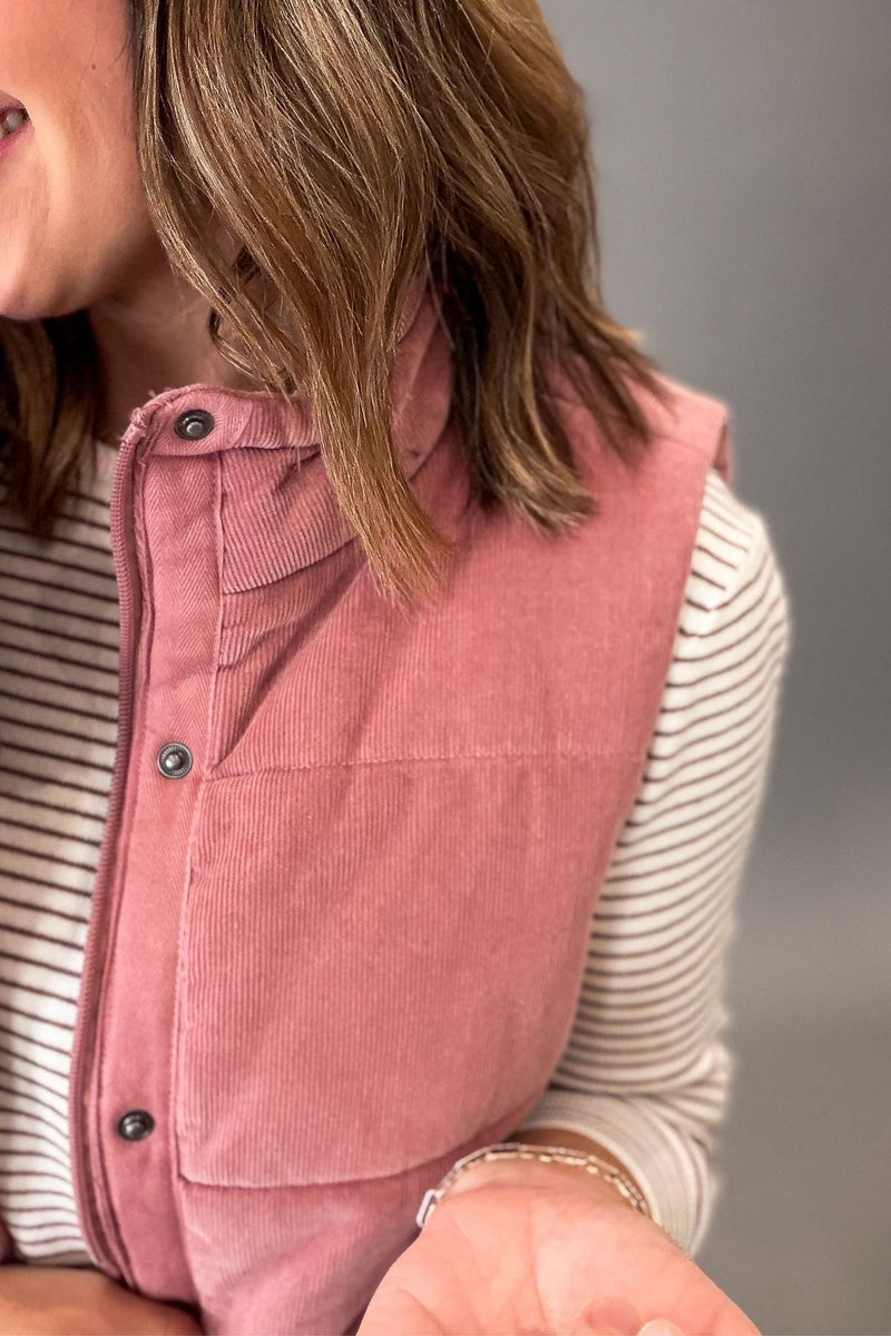 A close-up of a woman wearing a mauve corduroy vest over a mocha and white striped long-sleeve shirt, set against a plain gray background.