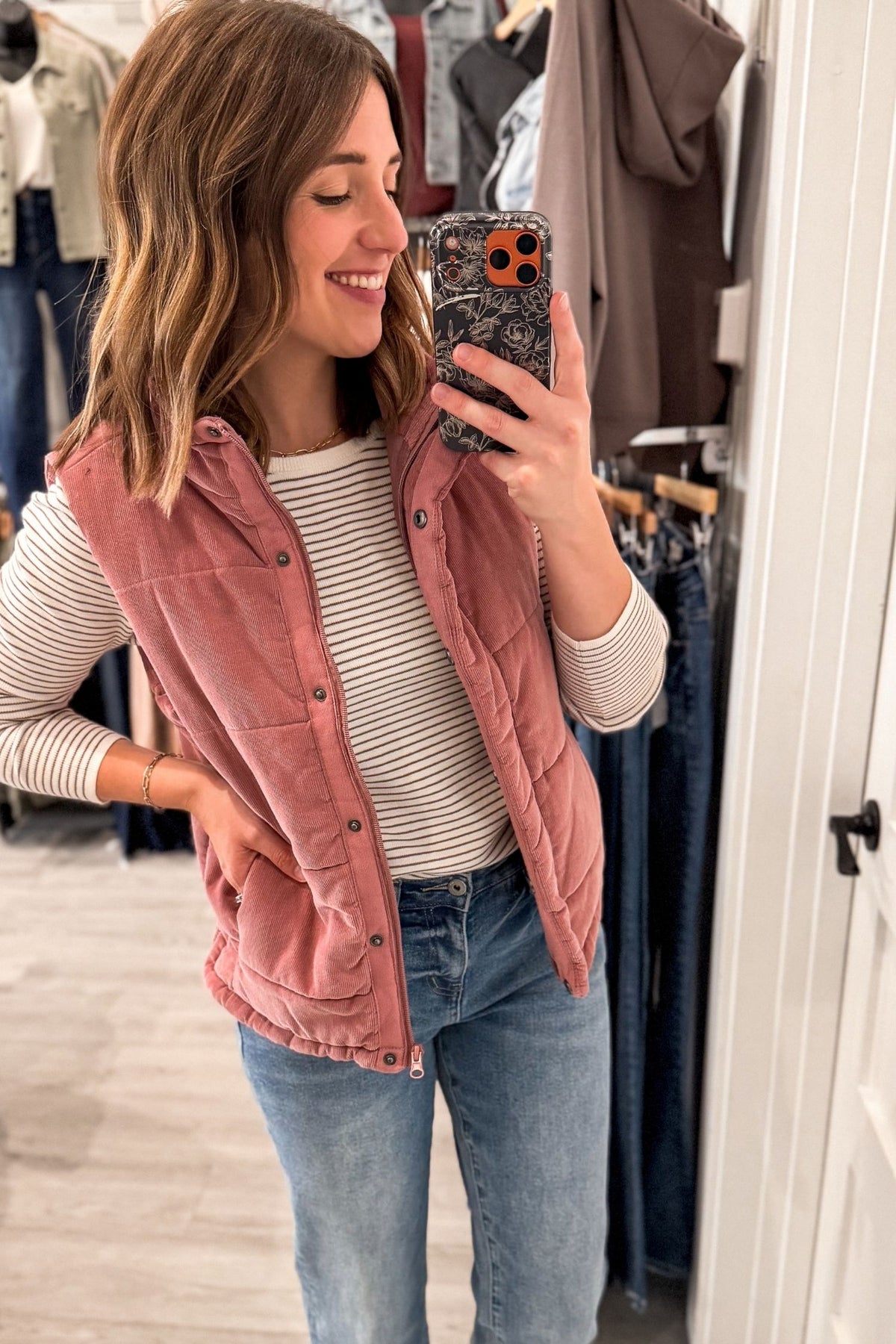 A woman takes a mirror selfie in a clothing store while wearing a mauve corduroy snap-front + zip vest over a ivory and mocha striped long-sleeve shirt and blue jeans.