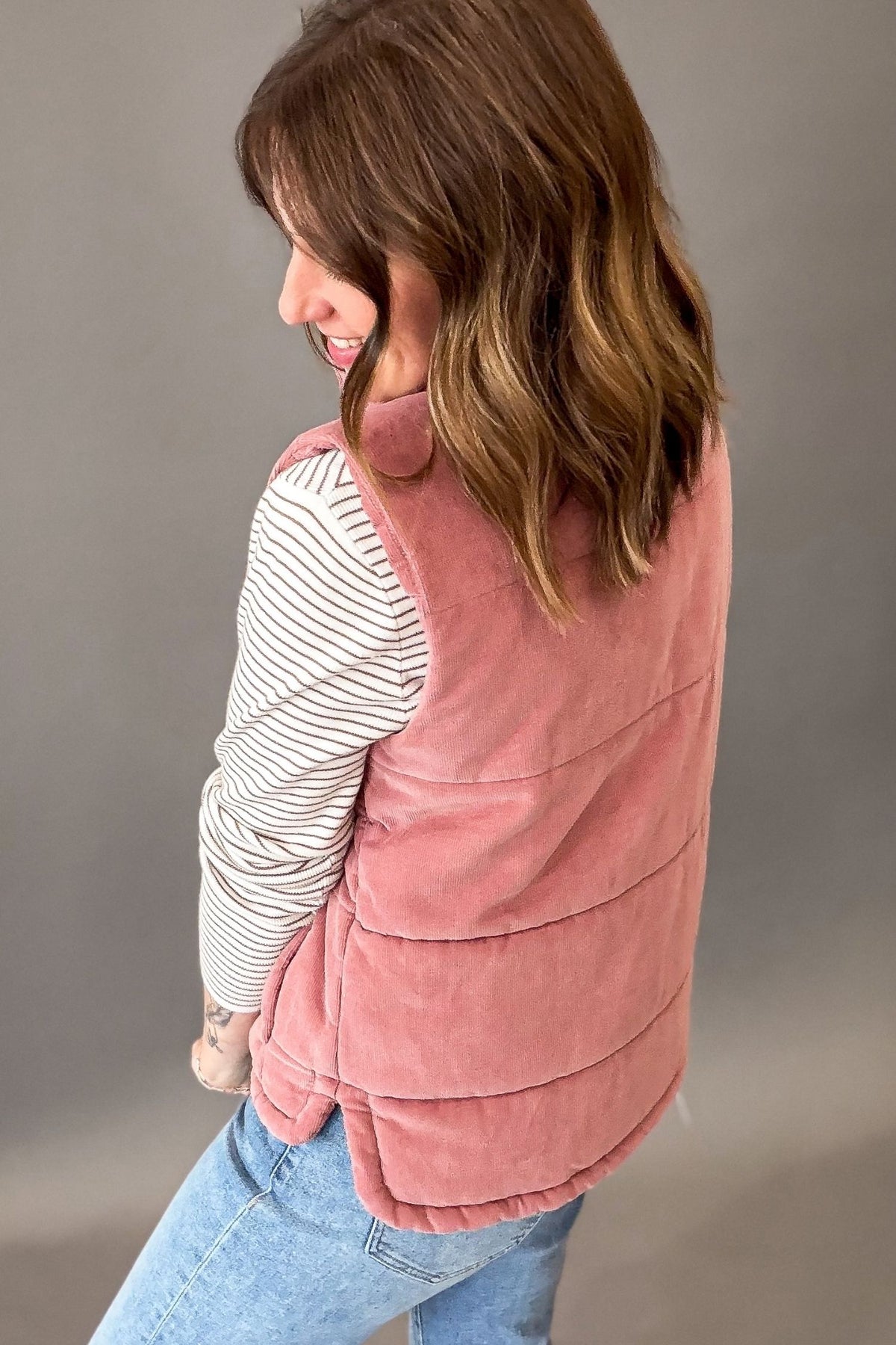 A woman shown from the back wearing a mauve corduroy puffer vest over a ivory long-sleeve striped shirt and blue jeans, standing against a neutral gray studio background.