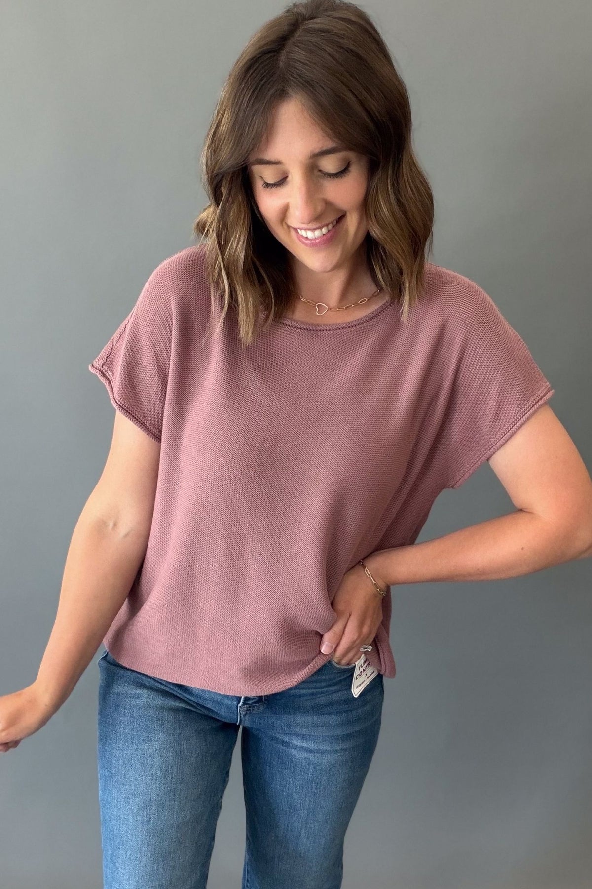 A woman wearing a mauve short-sleeve t-shirt and blue jeans, standing against a plain gray background.